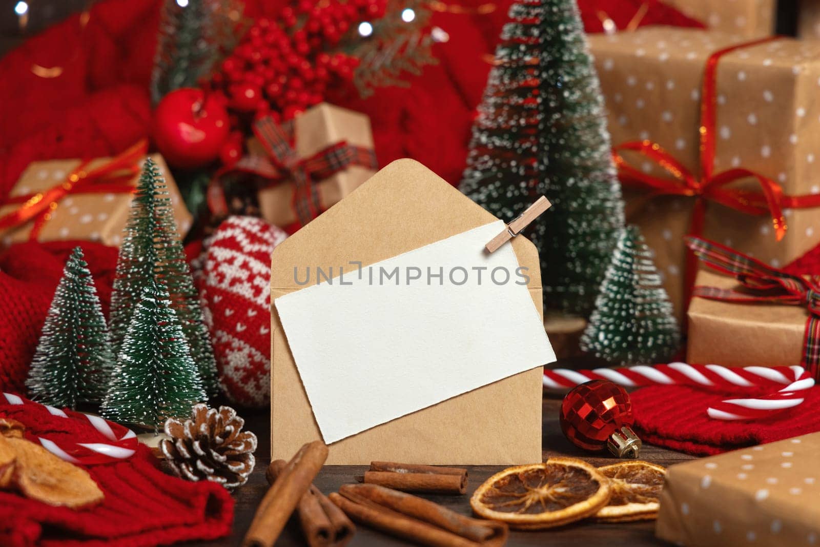 White postcard and craft envelope near Christmas decor, trees, spices and red knitted sweater on wooden table closeup, copy space. Winter atmospheric mock up, holiday greeting