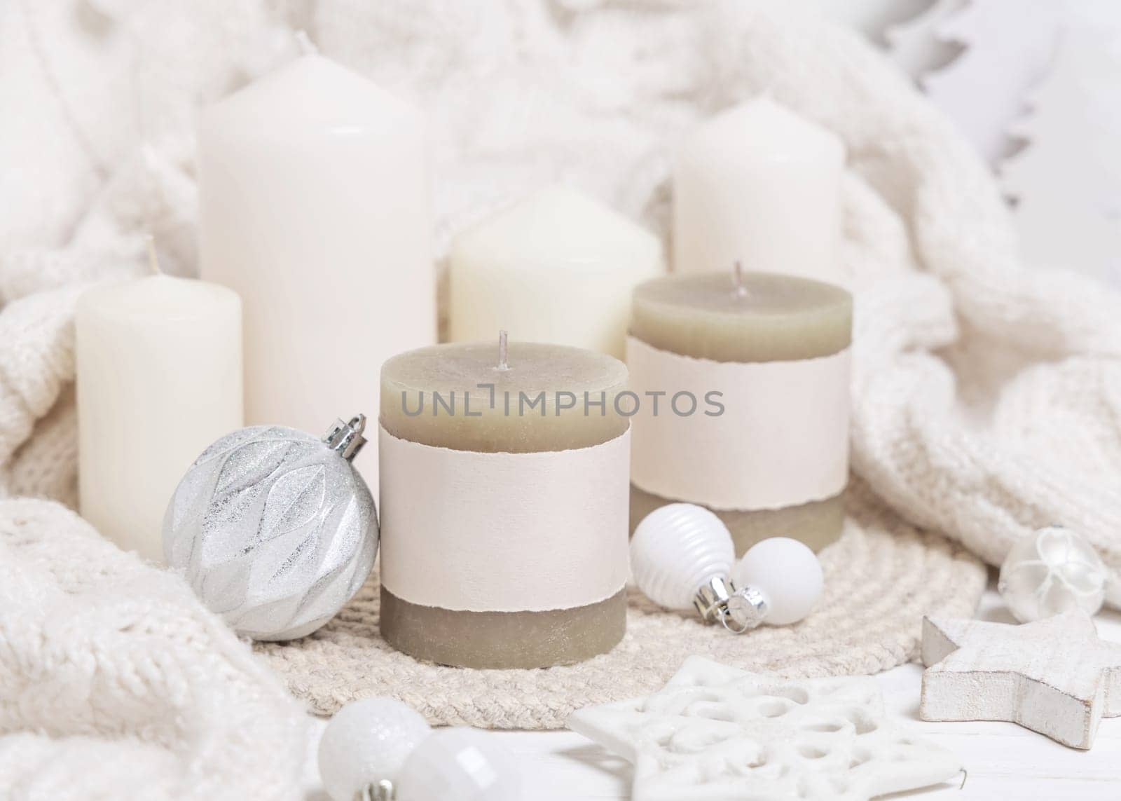Pillar candles with blank label near white knitted sweater, wooden Christmas decor and lights, copy space. Winter atmospheric mock up in rustic or scandinavian style