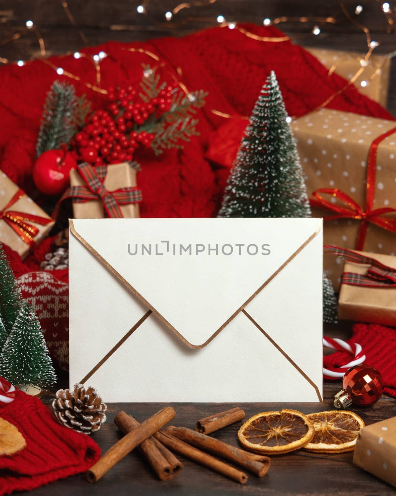 White golden trimmed envelope near Christmas decor, trees and red knitted sweater on wooden table closeup, place for text. Winter atmospheric mock up, family holiday time