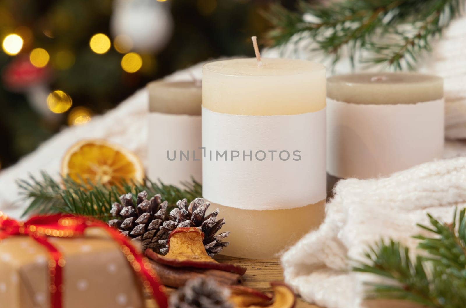Pillar candles with blank label near Christmas decor, dryed fruits and fir twigs close up against Christmas tree lights, copy space. Warm winter family atmospheric mock up, magical Holiday time