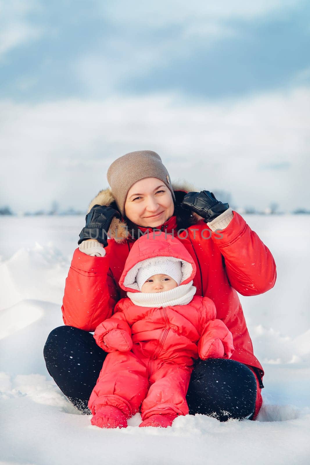 Happy mother and baby are sitting on the snow and enjoying a beautiful winter day