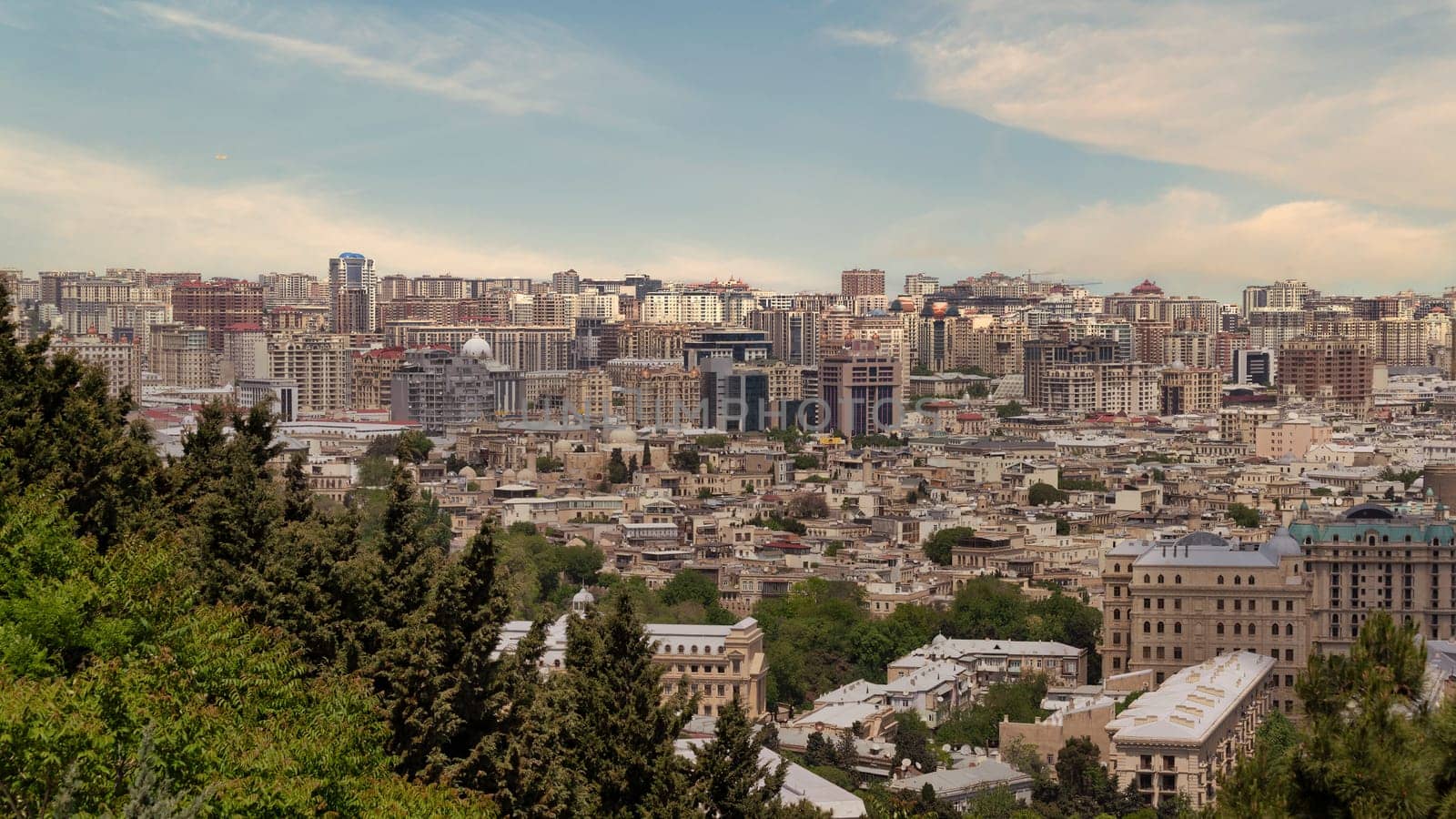 A panoramic view of Baku, Azerbaijan, showcases the contrast between modern skyscrapers and historic buildings in the foreground. Lush greenery envelops the hills, enhancing the scenic beauty