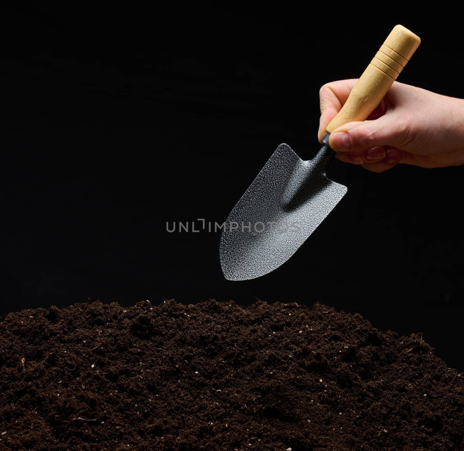 A woman's hand holds a small garden trowel over a pile of damp, loose soil, the beginning of farming