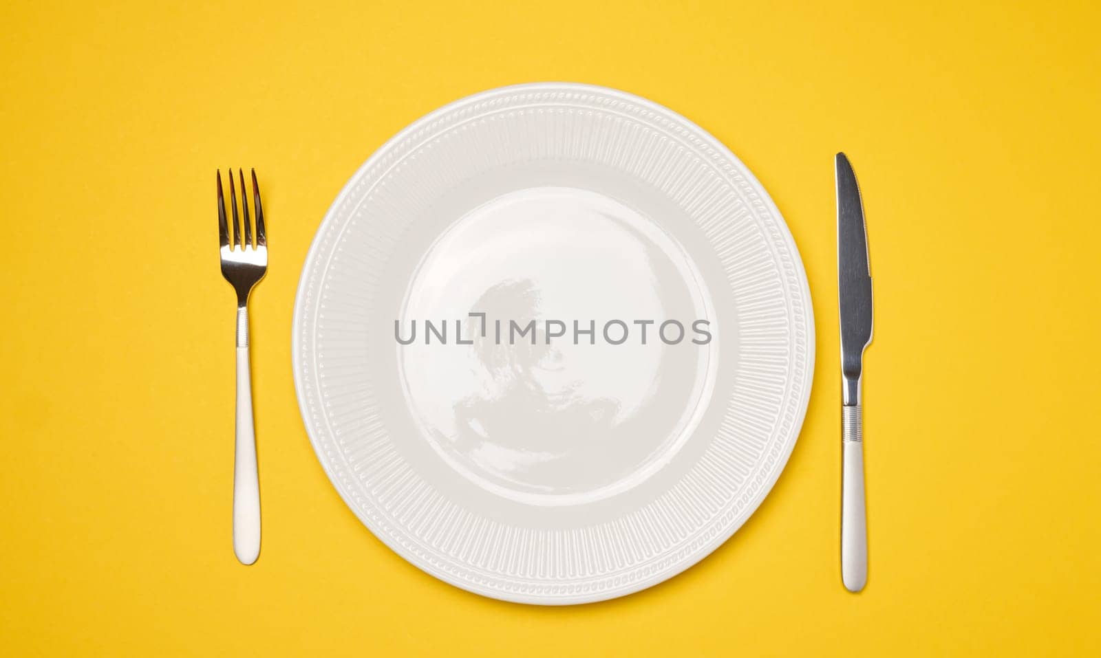 Round white empty ceramic plate and knife with fork on yellow background, table setting. Top view