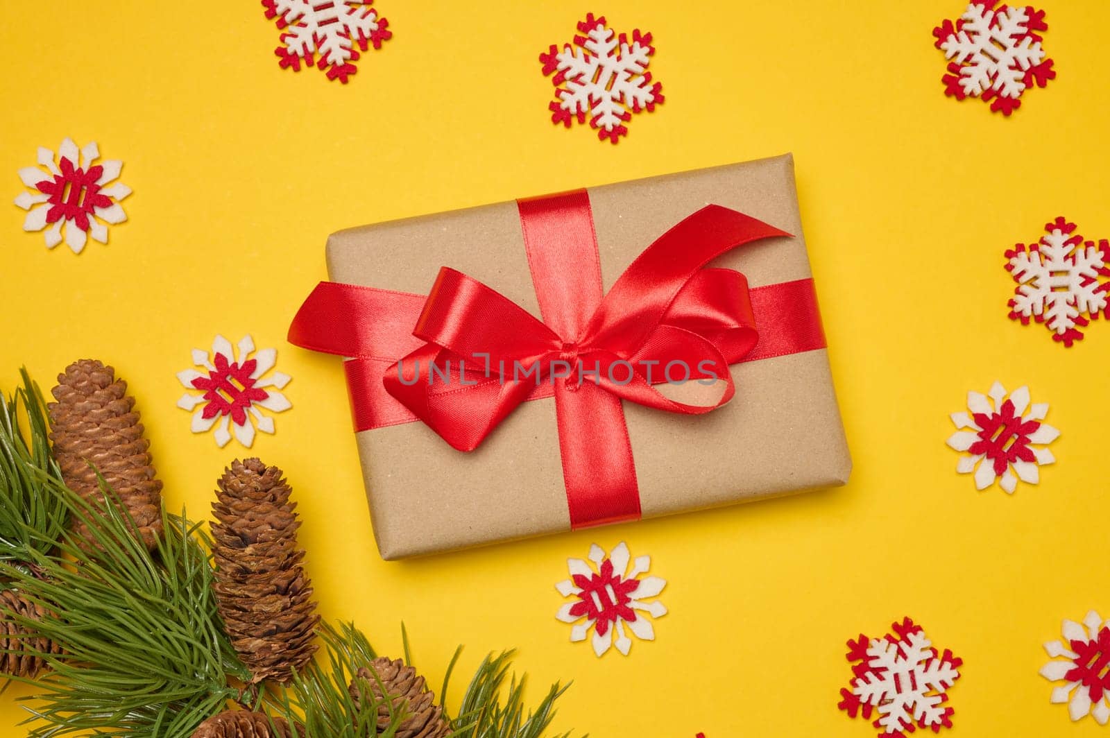 Gift box with red satin ribbon and bow on a yellow background. Holiday background. There is a pine branch with cones nearby