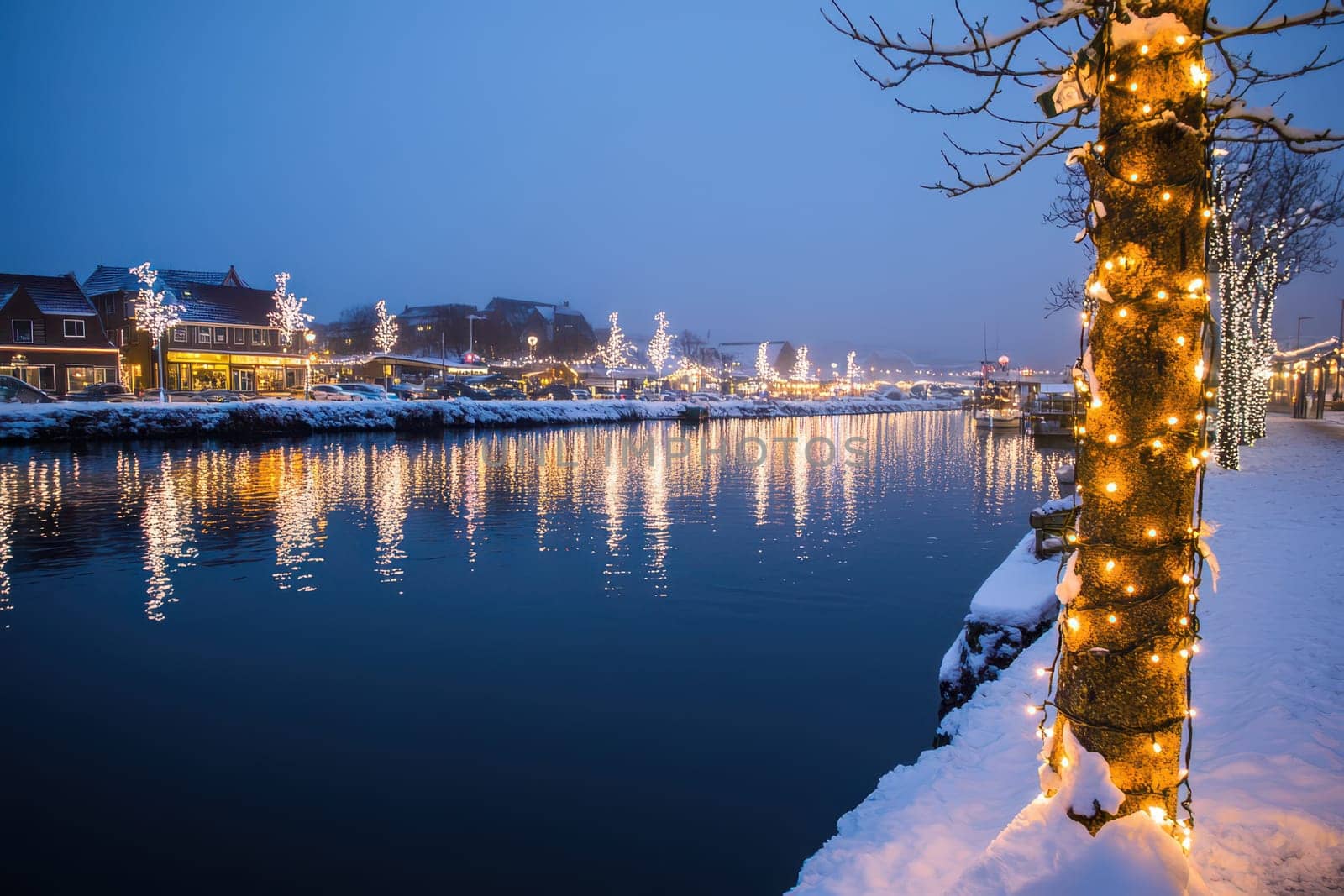 A serene river scene showcases festive lights on trees along the bank surrounded by snow covered buildings during a peaceful winter evening.
