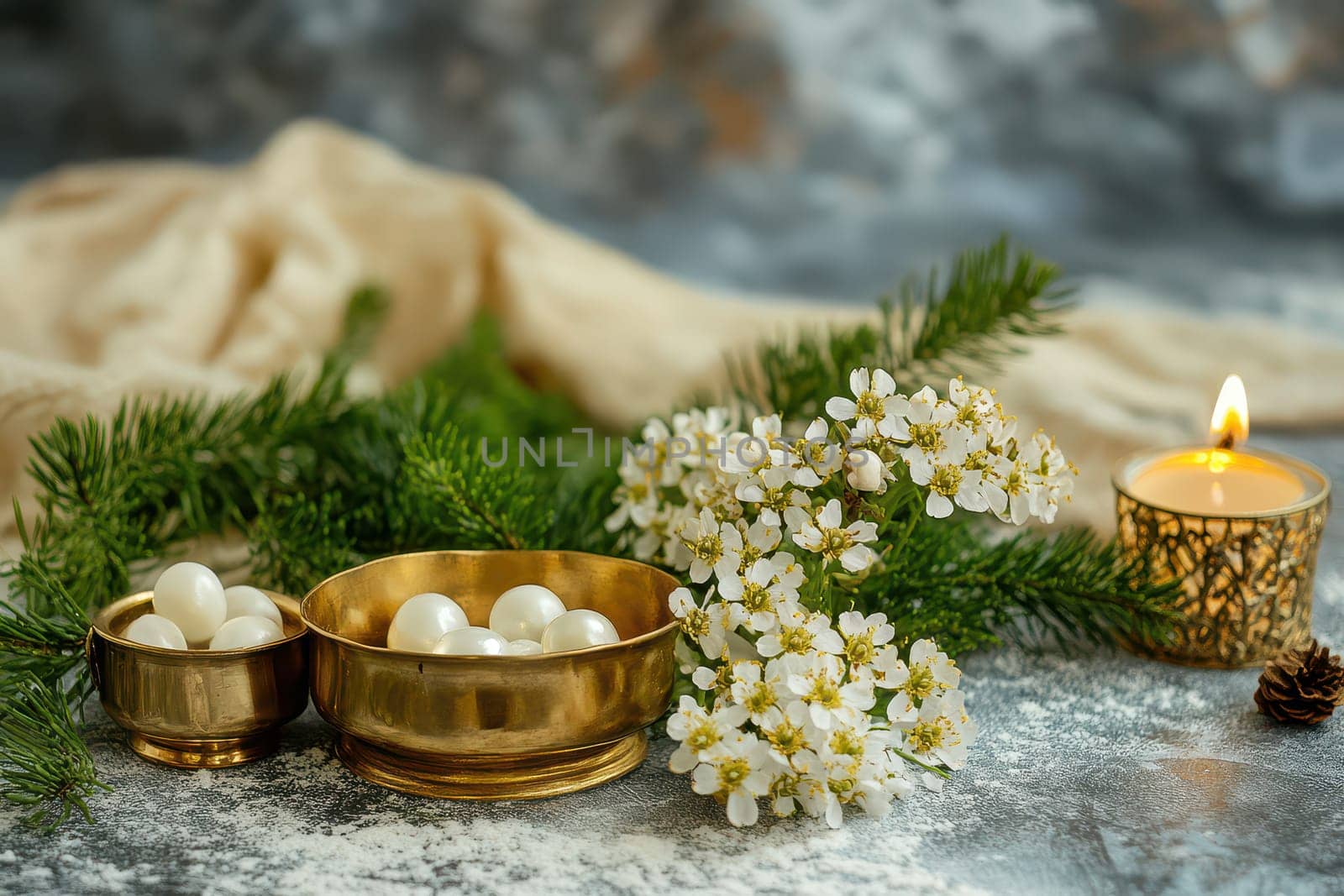 Delicate white flowers and golden bowls with pearls are arranged on a textured surface accompanied by a glowing candle and greenery.