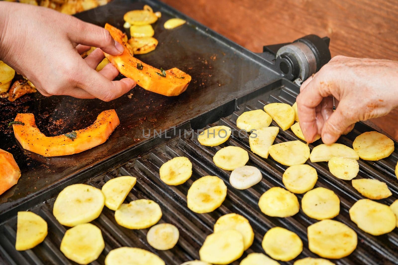 Orange butternut squash pieces grilled on electric grill, detail on woman hands moving smoking vegetables, blurred potato chips in front