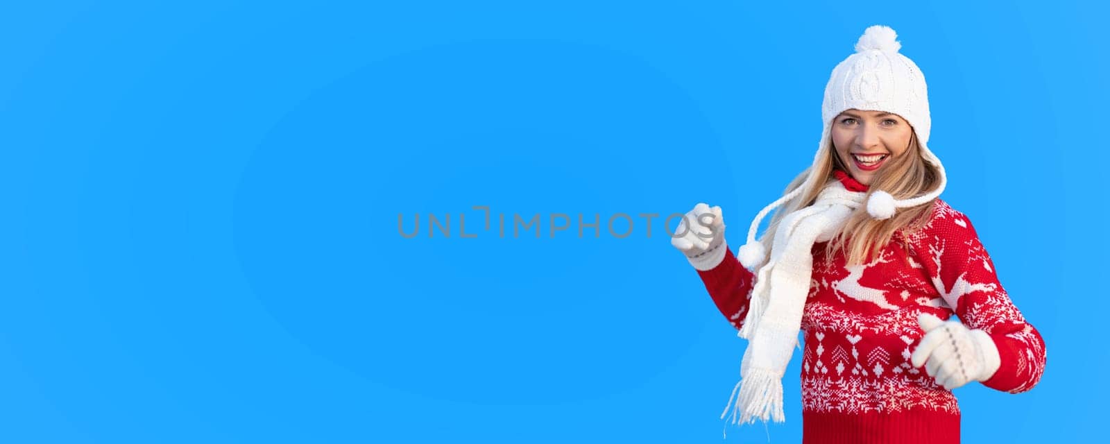 Young woman in red winter pullover jumper, white gloves, ski hat and muffetee, smilling happily, isolated on blue background (replace with your image or add text to copy space)