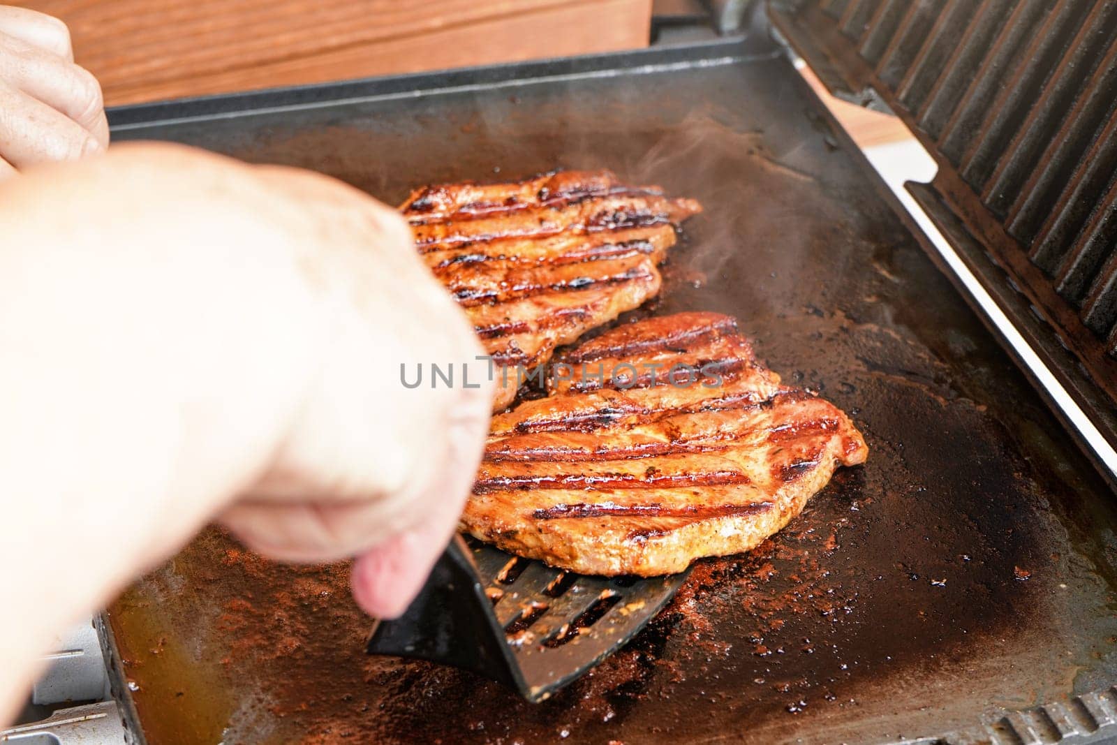 Two pork cutlet steak grilled on electric grill, blurred woman hand holding plastic spatula, about to turn one piece of meat