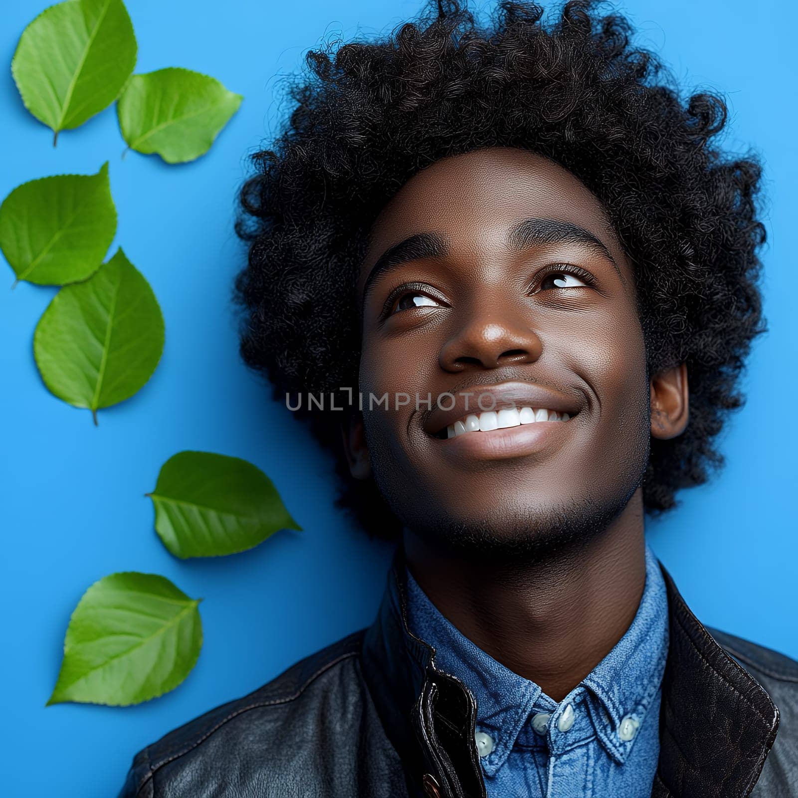 Stylish young man in leather jacket and denim shirt against blue background with green leaves, natural cheerful portrait with bright smile.