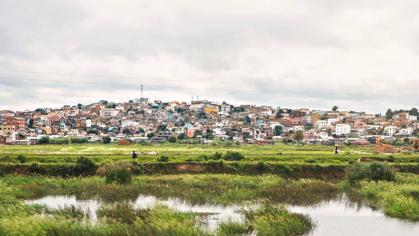Antananarivo, Madagascar - April 24, 2019: Typical scenery during overcast day near Madagascar capital, houses on small hills background, with people working at wet rice fields in foreground