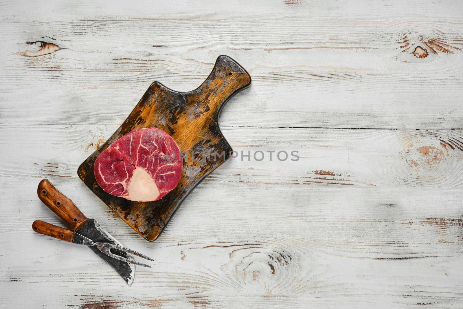 A raw beef steak on a wooden cutting board. Butcher knife and fork are placed below the cutting board on a white wooden tabletop
