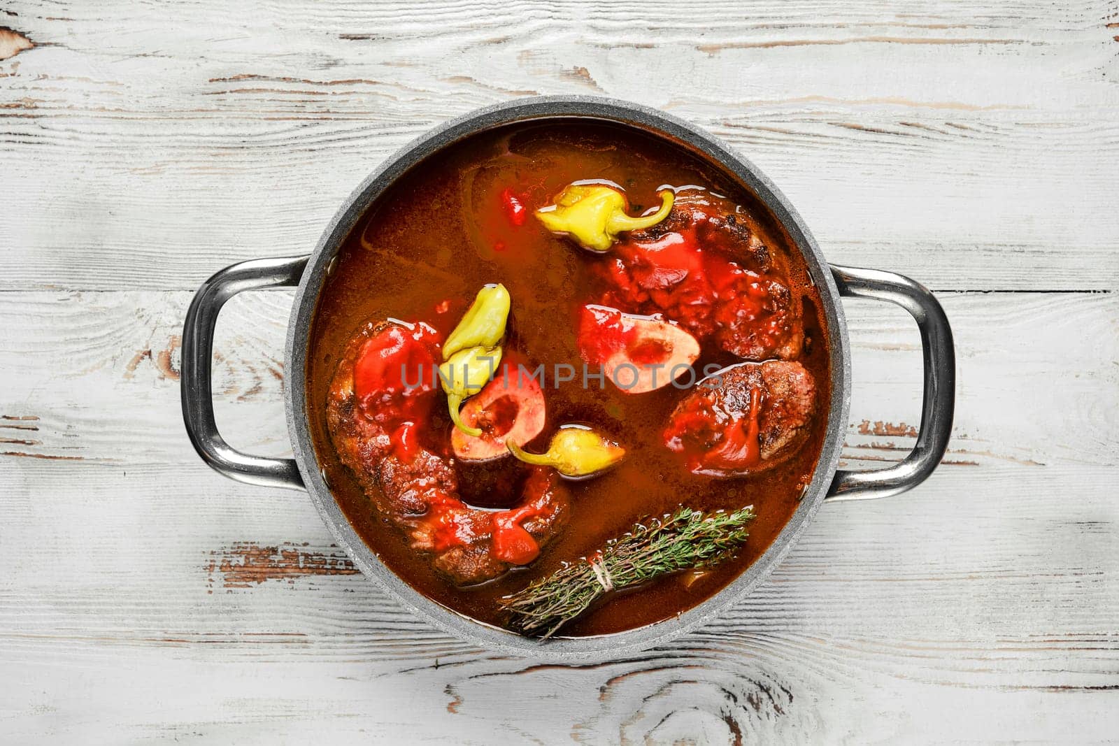 A pot of beef stew is on a white wood table. The stew is simmering and contains large pieces of beef, peppers, and herbs. The pot has two handles.