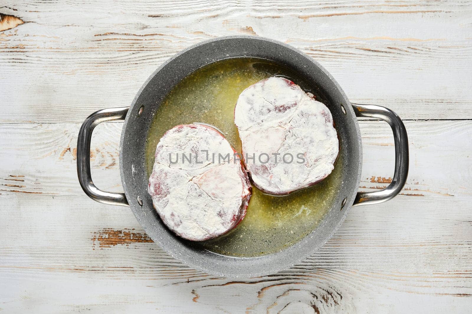 Overhead view of two floured steaks are sizzling in a pan with oil. The pan is on a white wooden tabletop