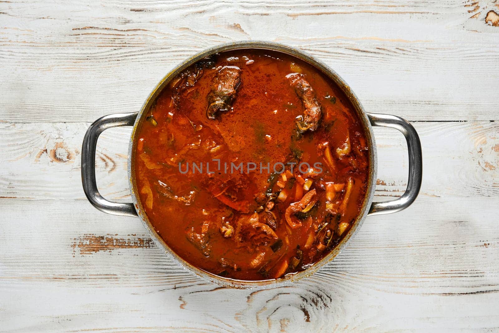 Overhead view of a silver pot with two handles on a white wooden table. The pot is filled with a rich beef stew, filled with chunks of meat and vegetables.