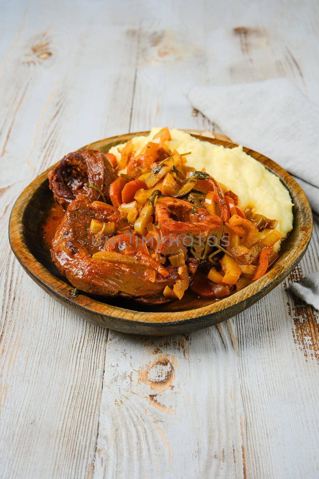 Osso buco, a traditional Italian dish, is served on a wooden plate with mashed potatoes, onions, peppers, and a rich gravy.