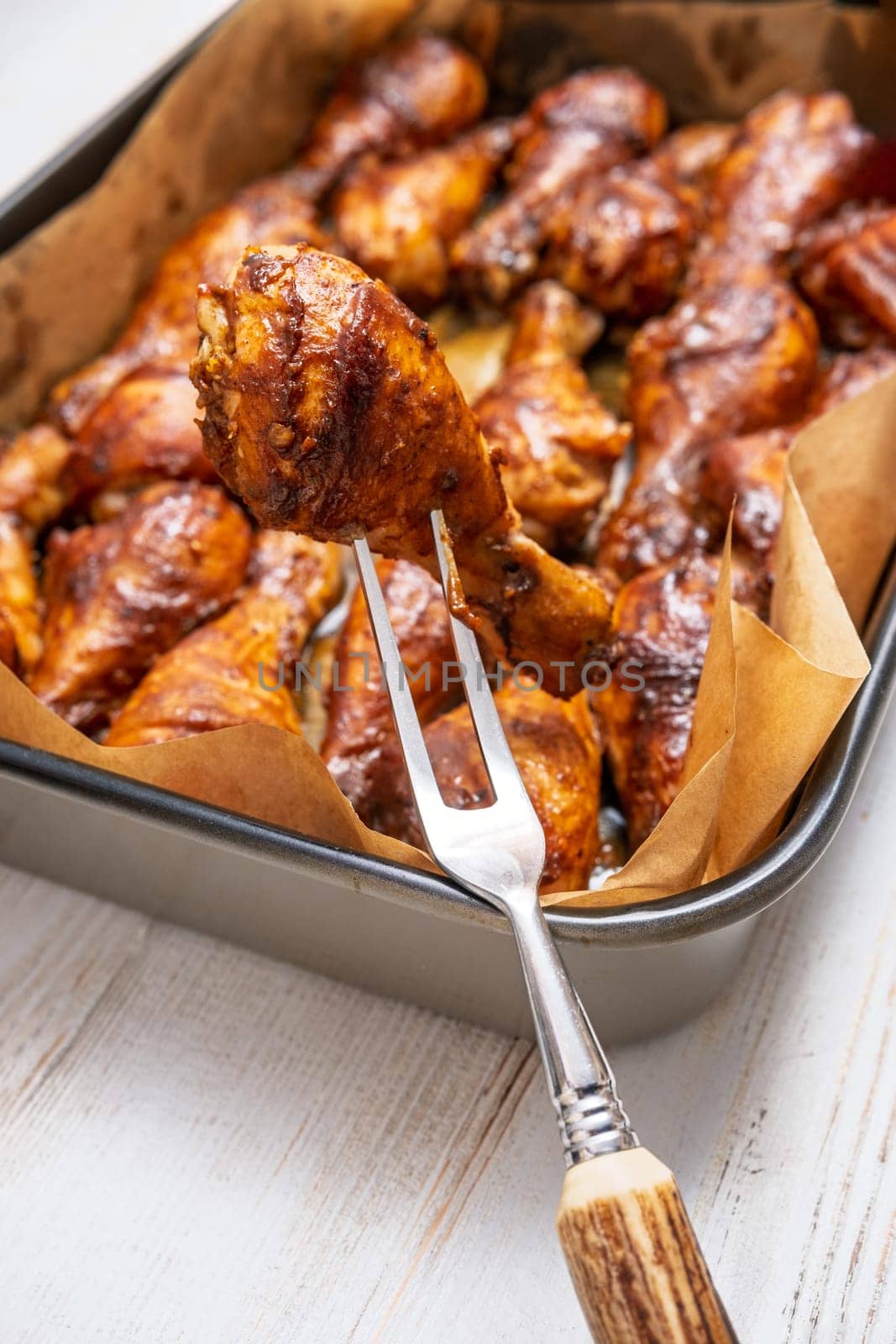 A fork lifts a perfectly cooked chicken drumstick from a baking tray with parchment paper. The succulent pieces are glazed to a rich brown, showcasing an appetizing grilled appearance.