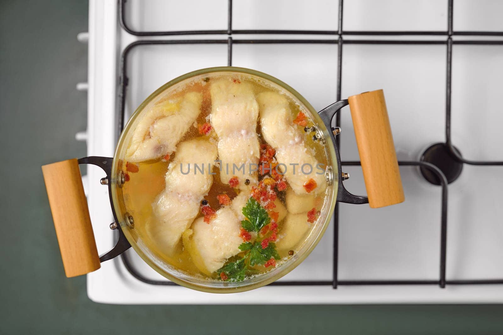 A pot of white fish fillets simmers slowly in clear broth, garnished with herbs and spices. The scene captures a warm, inviting kitchen atmosphere, emphasizing the home-cooked meal preparation.