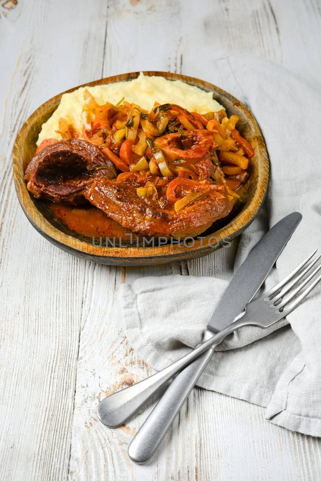 A wooden plate of osso buco served with mashed potatoes, cooked in a tomato and onion sauce with herbs. A silver knife and fork are placed beside a linen napkin on a white wooden table.