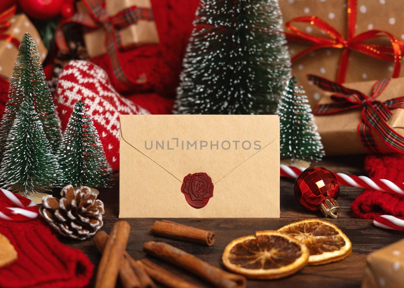 Beige sealed envelope near Christmas decor, trees and red knitted sweater on wooden table closeup, place for text. Winter mock up for greeting card or invitation