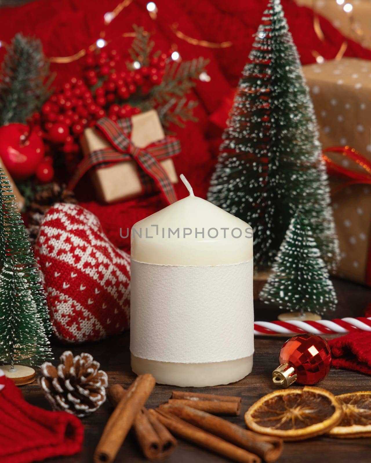 Pillar candle with blank label near red knitted sweater and Christmas decor against lights and wrapped presents, copy space. Warm winter family atmospheric mock up, Holiday time