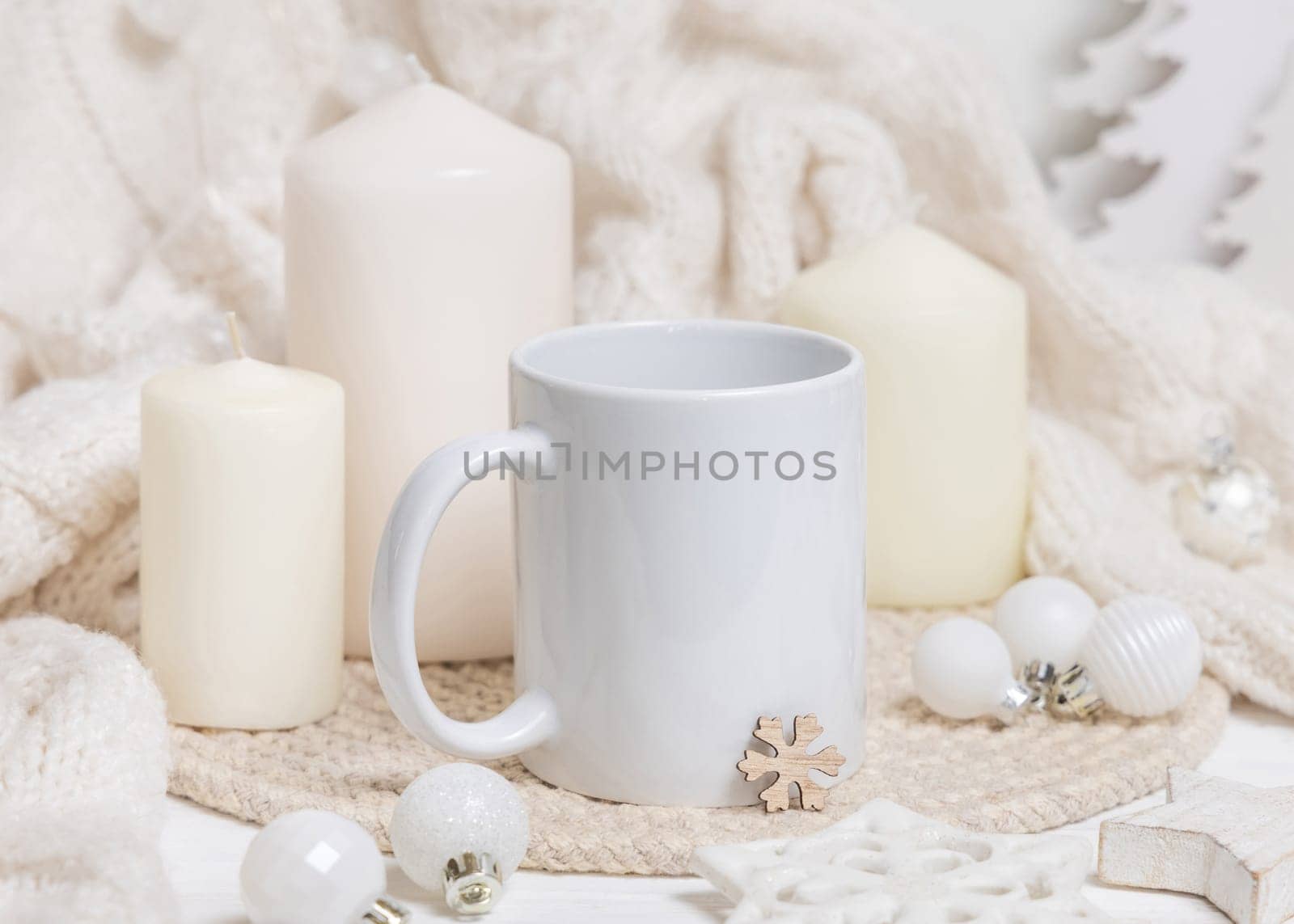White blank coffee mug near candles, white knitted sweater and Christmas decor, copy space. Winter atmospheric mockup in rustic or scandinavian style