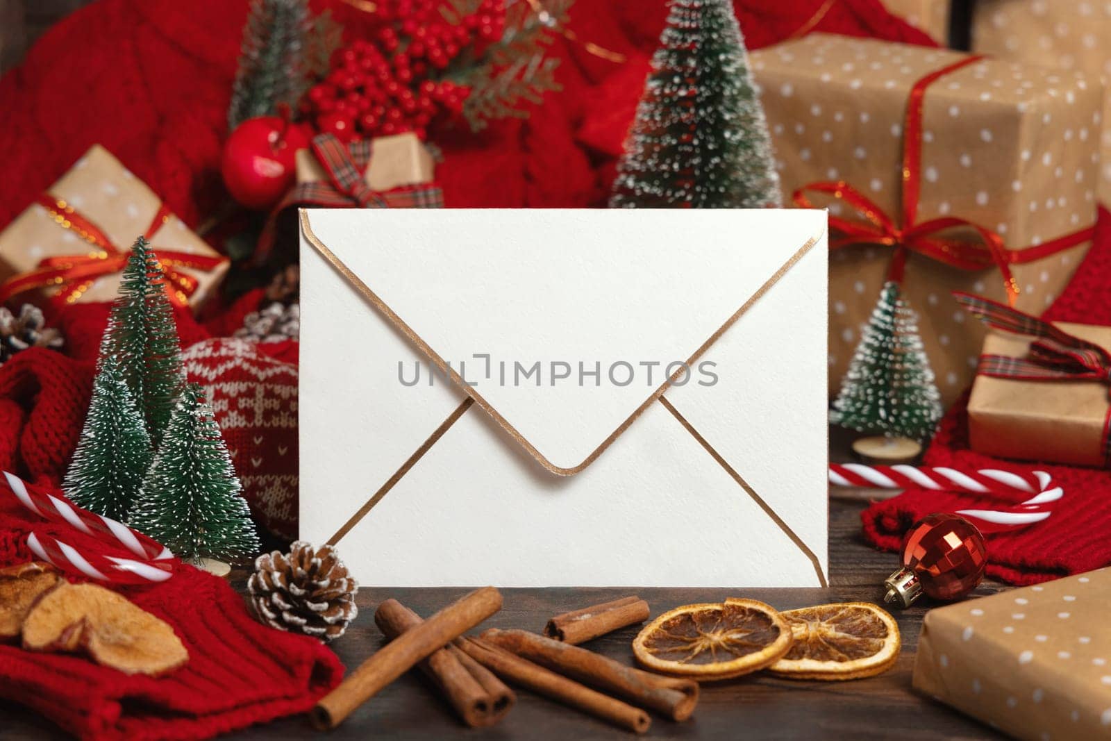 White golden trimmed envelope near Christmas decor, trees and red knitted sweater on wooden table closeup, place for text. Winter atmospheric mock up, family holiday time