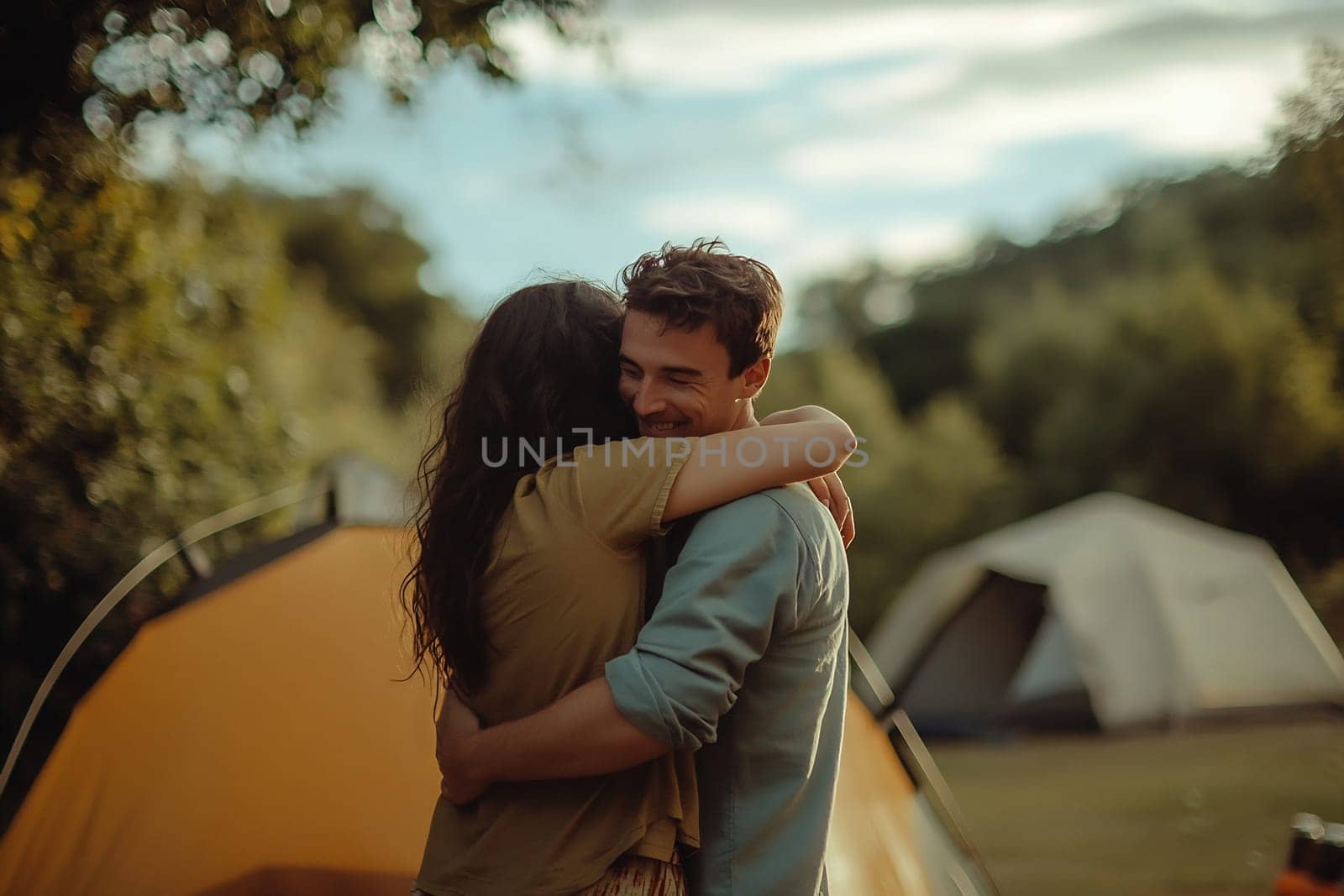 A joyful couple shares a warm embrace in nature, surrounded by camping tents and a serene landscape during golden hour.