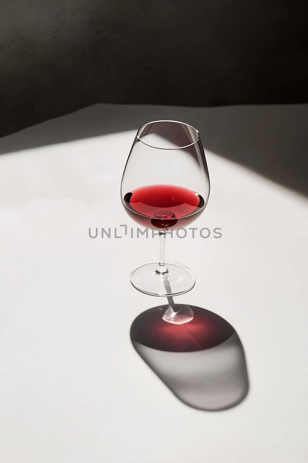A wine glass filled with red wine sits on a white surface, illuminated by soft light that creates a delicate shadow.
