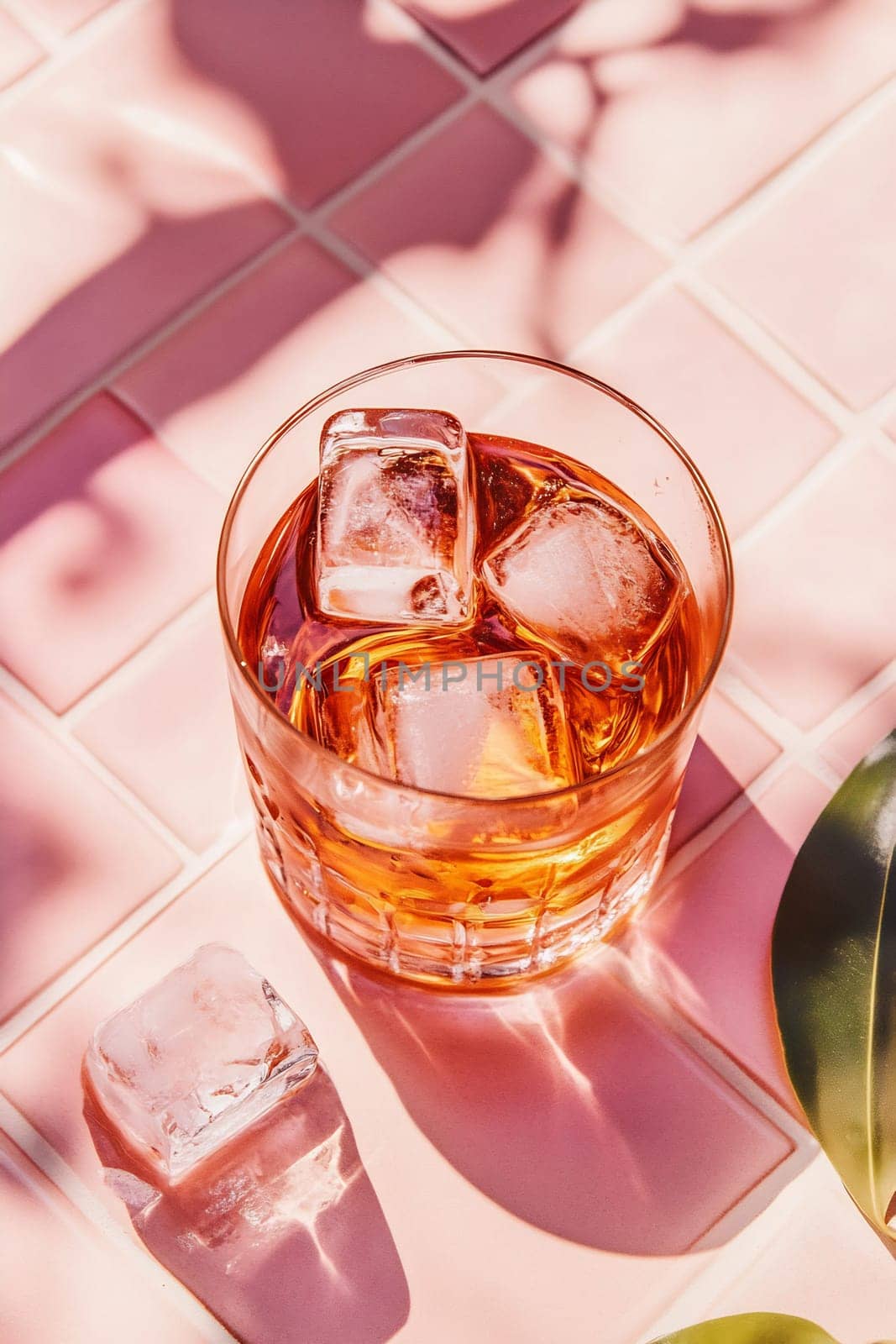 A glass filled with a chilled beverage and ice sits on a pink tiled surface, basking in warm sunlight.