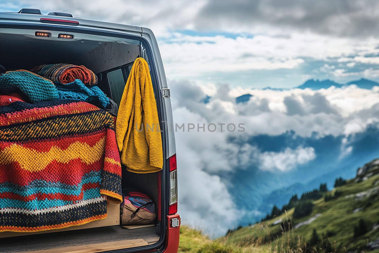 A colorful blanket and shawl hang from a van in a scenic mountain landscape with clouds below and peaks above, showcasing outdoor adventure.