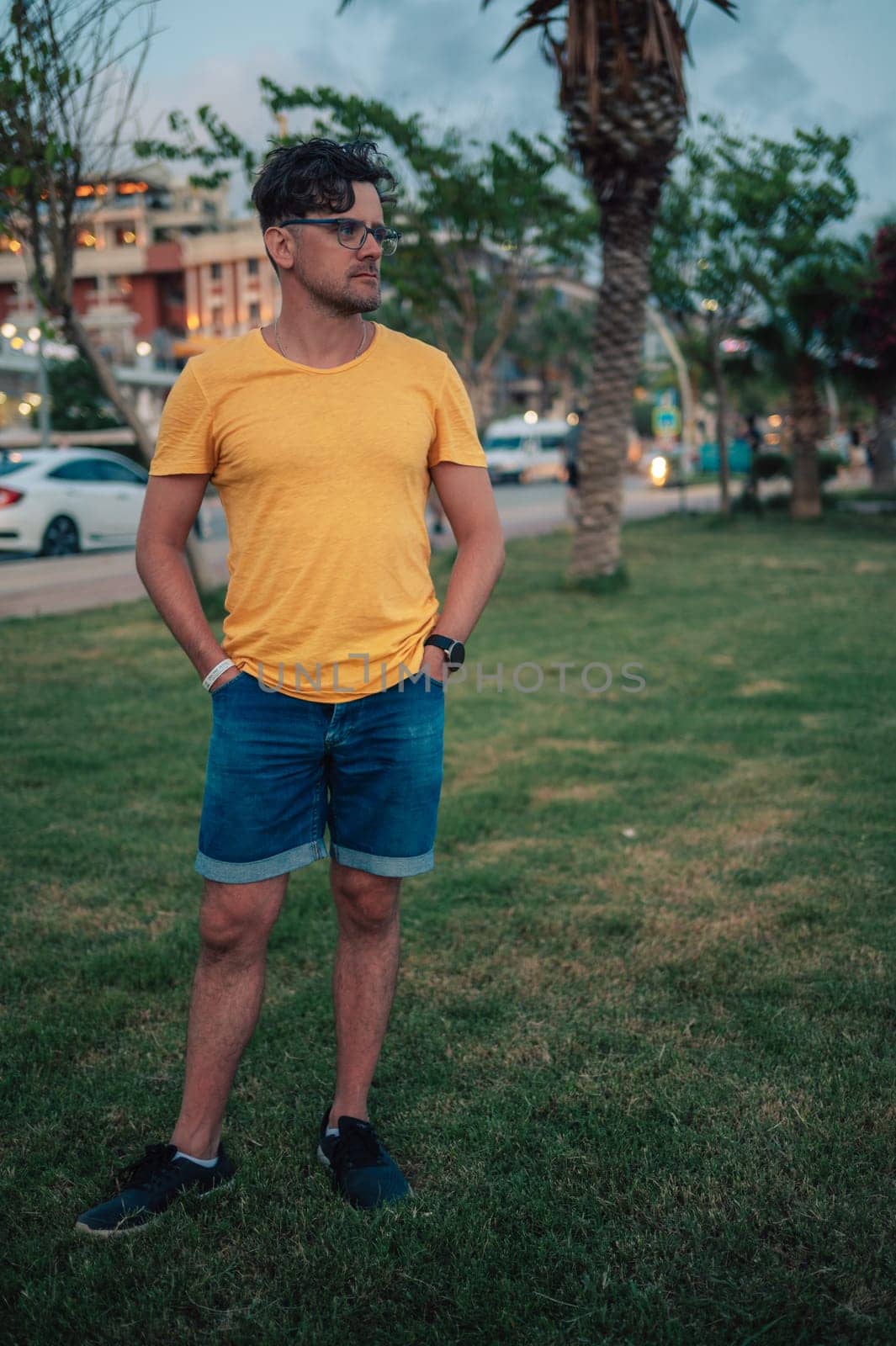 Man wearing a yellow shirt and glasses standing outdoors at dusk, with blurred city lights and greenery in the background.