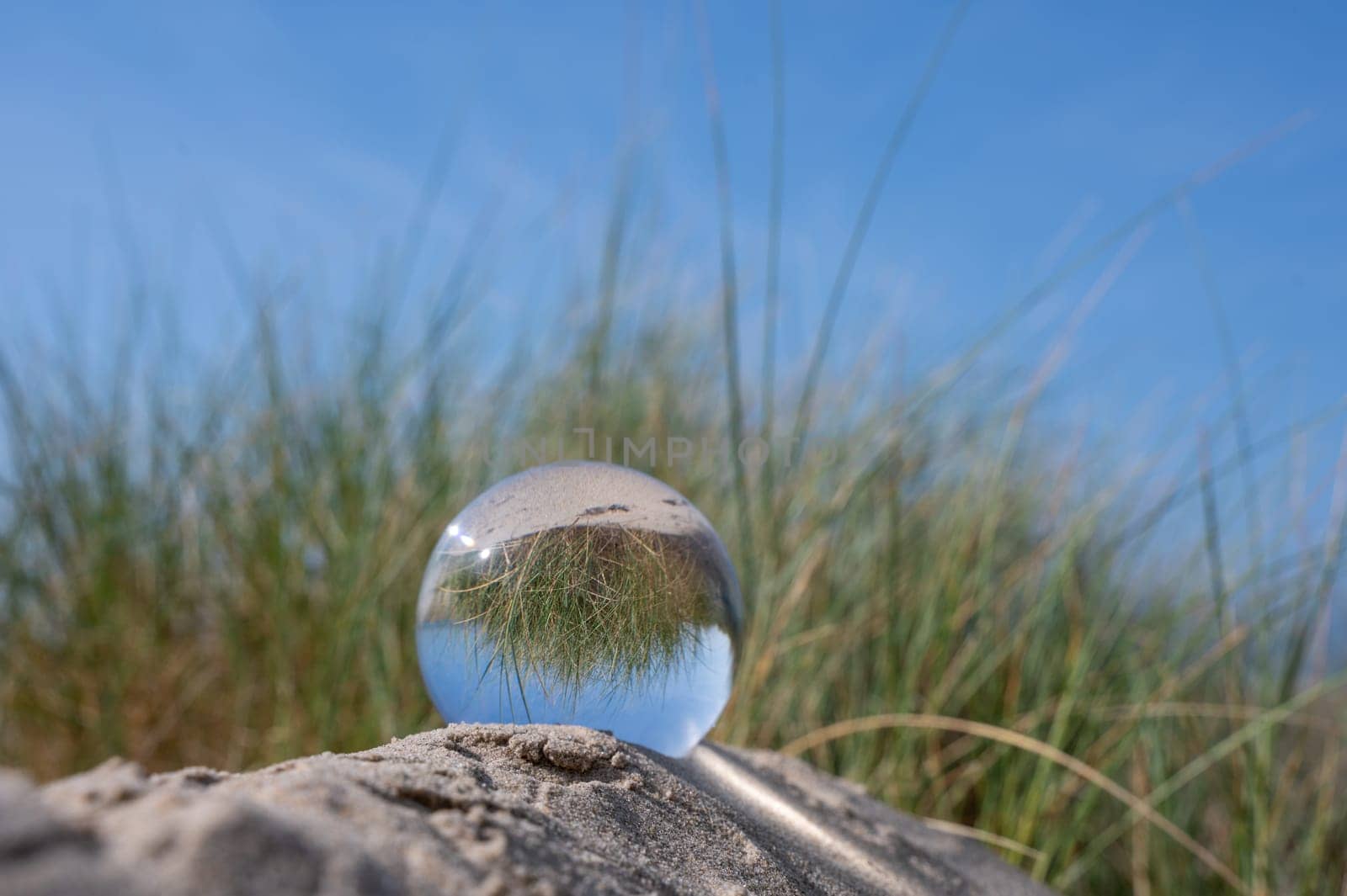 A glass ball lies in the sand of a dune by the sea, the dune grass and the blue sky are reflected in the ball