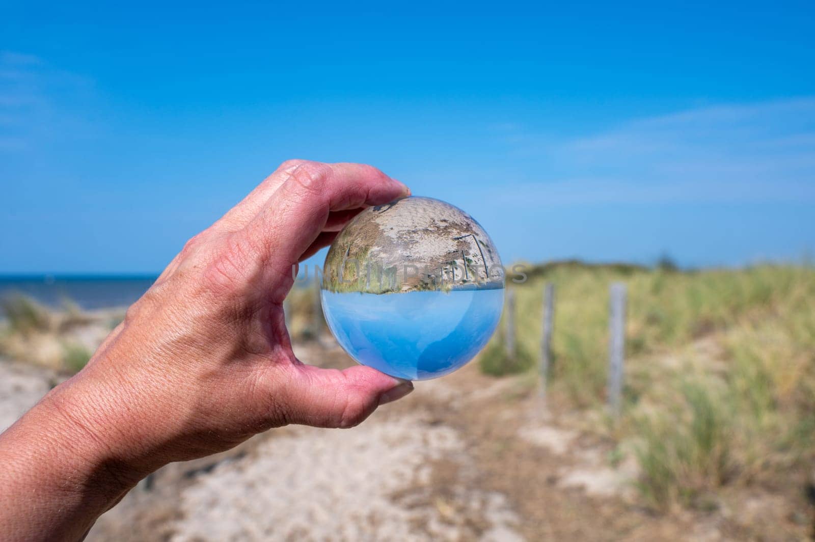 A human hand holds a glass ball, in the ball a sand dune, a footpath, blue sky and the sea are reflected