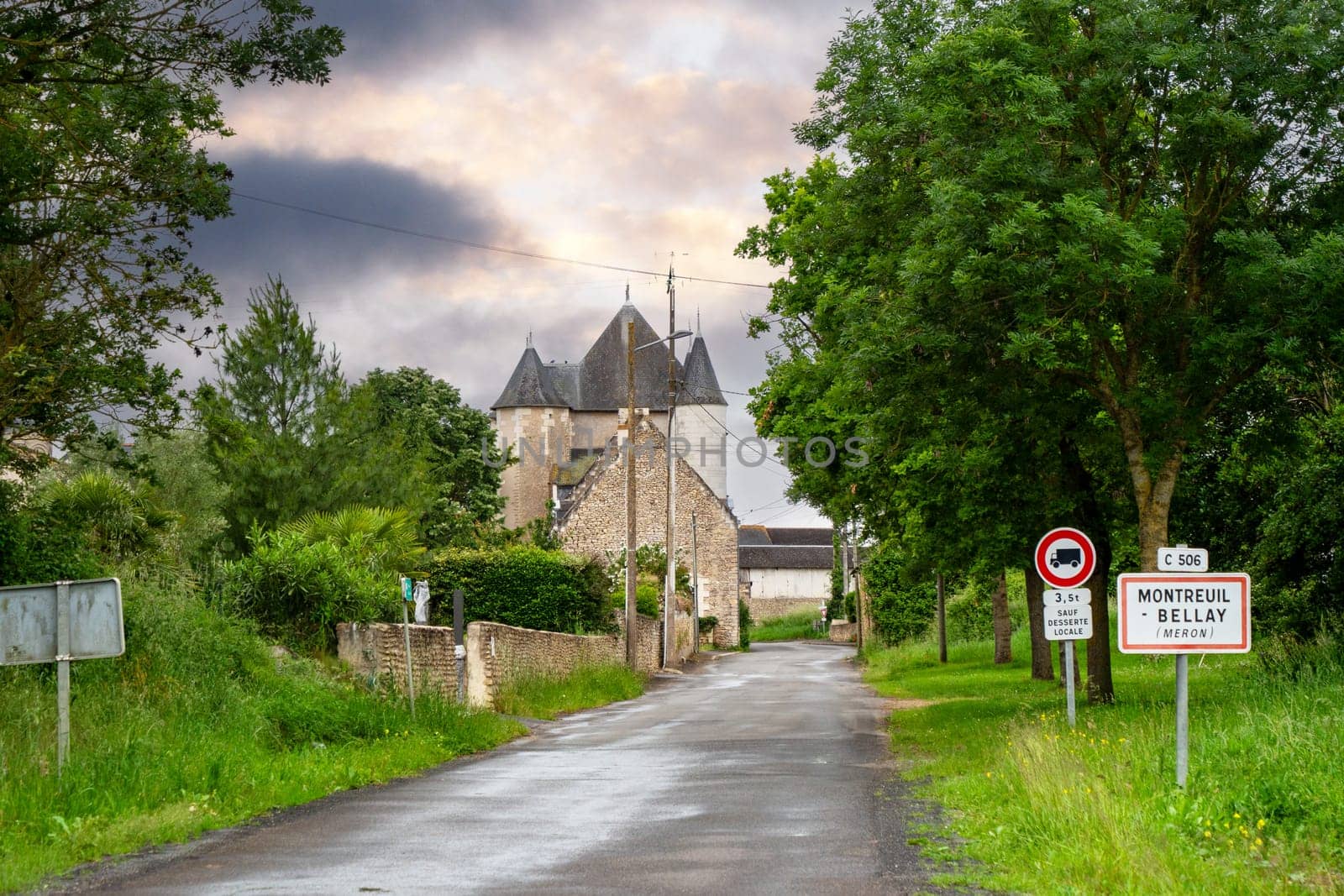 A road with a house and a sign that says Montreuil Bellay. The sky is cloudy and the trees are green, France