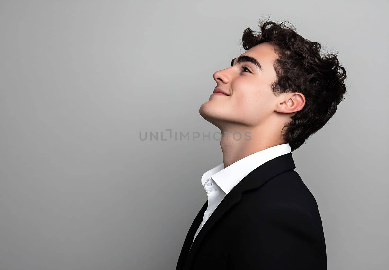 A young man dressed in a suit smiles softly while looking upward, exuding a sense of confidence and optimism against a plain background.