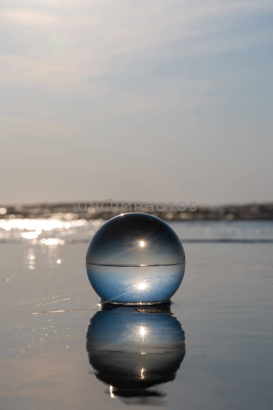 A glass ball lies in the waves on the sandy beach, the sea and the setting sun are reflected in the ball