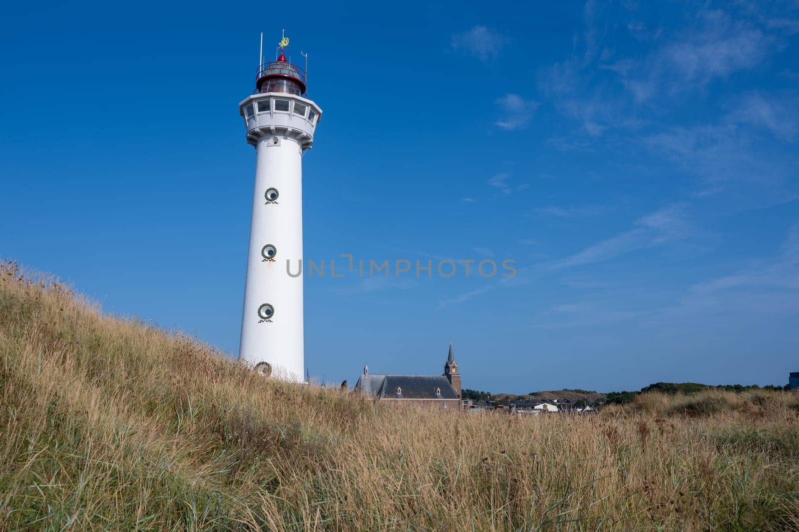 The approx. 28 m high white lighthouse J.C.J. van Speijk in Egmond aan Zee, North Holland, Netherlands with the Catholic St. Agnes Church, beach grass and sand dunes