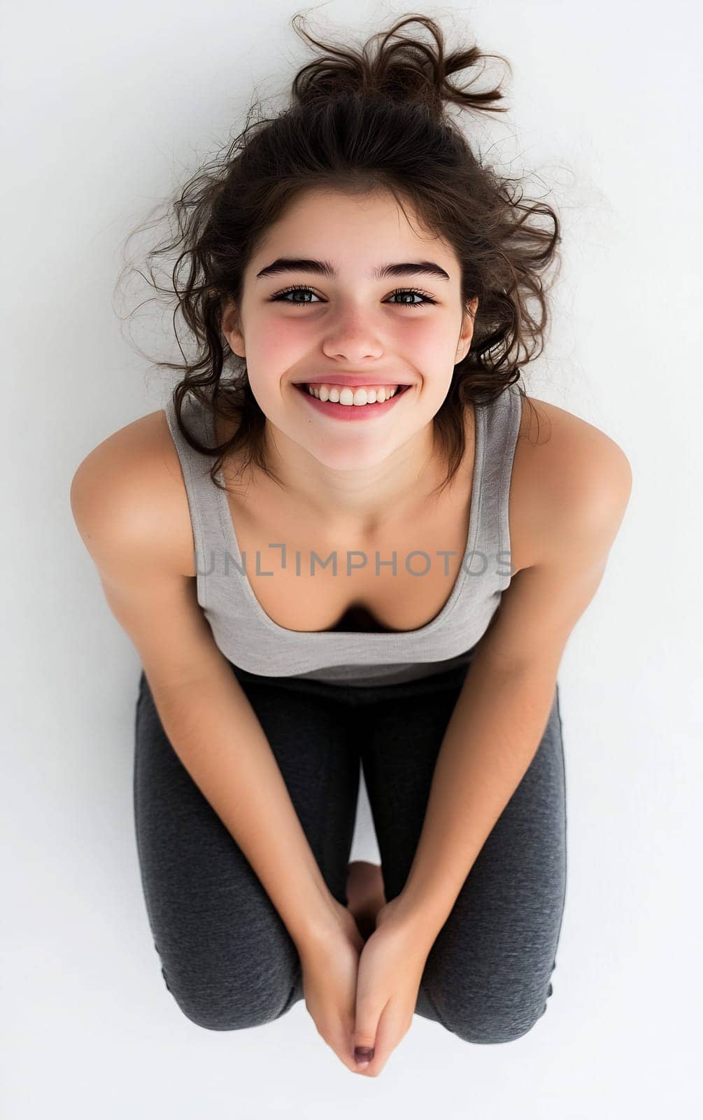 A joyful young woman sits on the floor with a bright smile, her hair in a messy bun against a clean background.