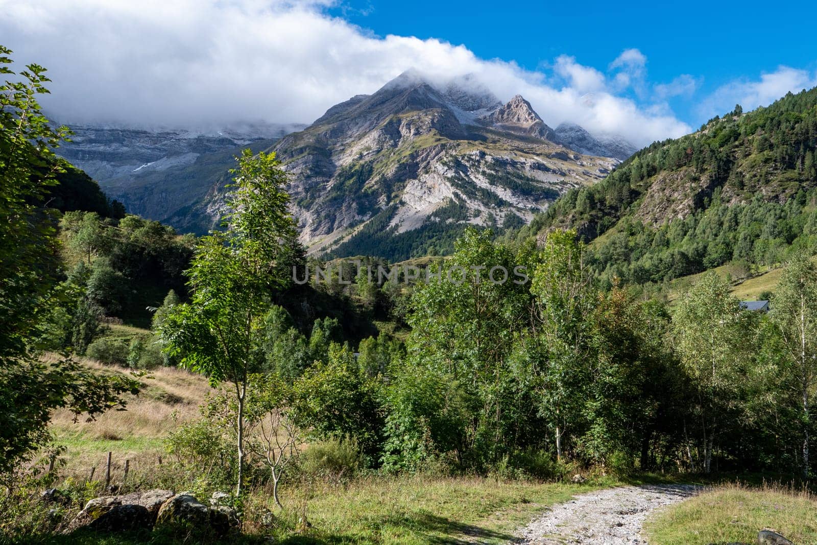 A mountain range with a cloudy sky in the background. The trees are green and the sky is blue. Cirque de Gavarnie, France