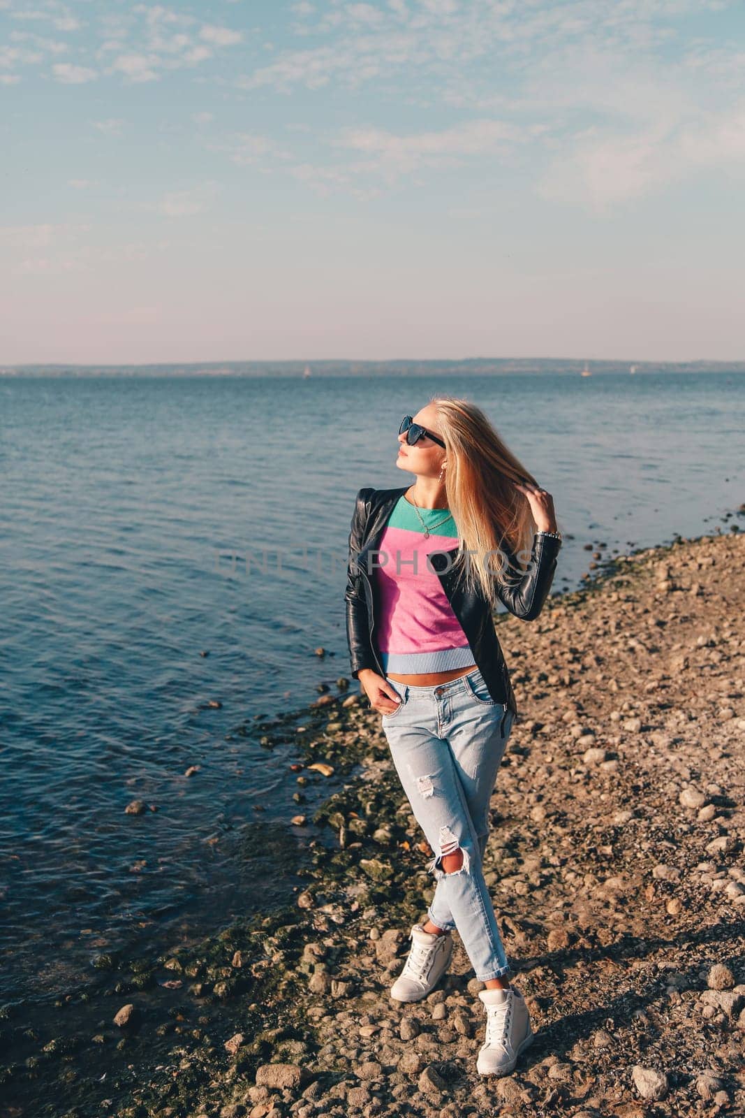 Fashion model with sunglasses, ripped jeans, pink top and black leather jacket posing on a pebbly beach by the sea or lake