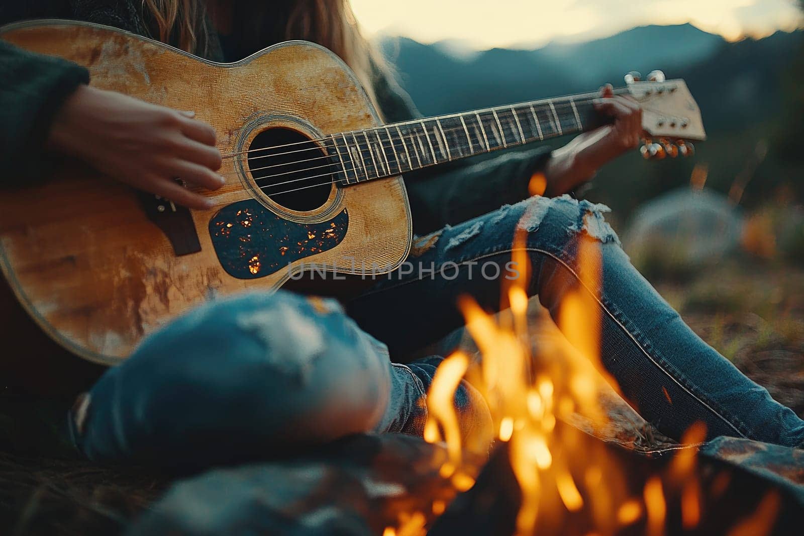 Young woman is playing acoustic guitar while sitting near a campfire at dusk