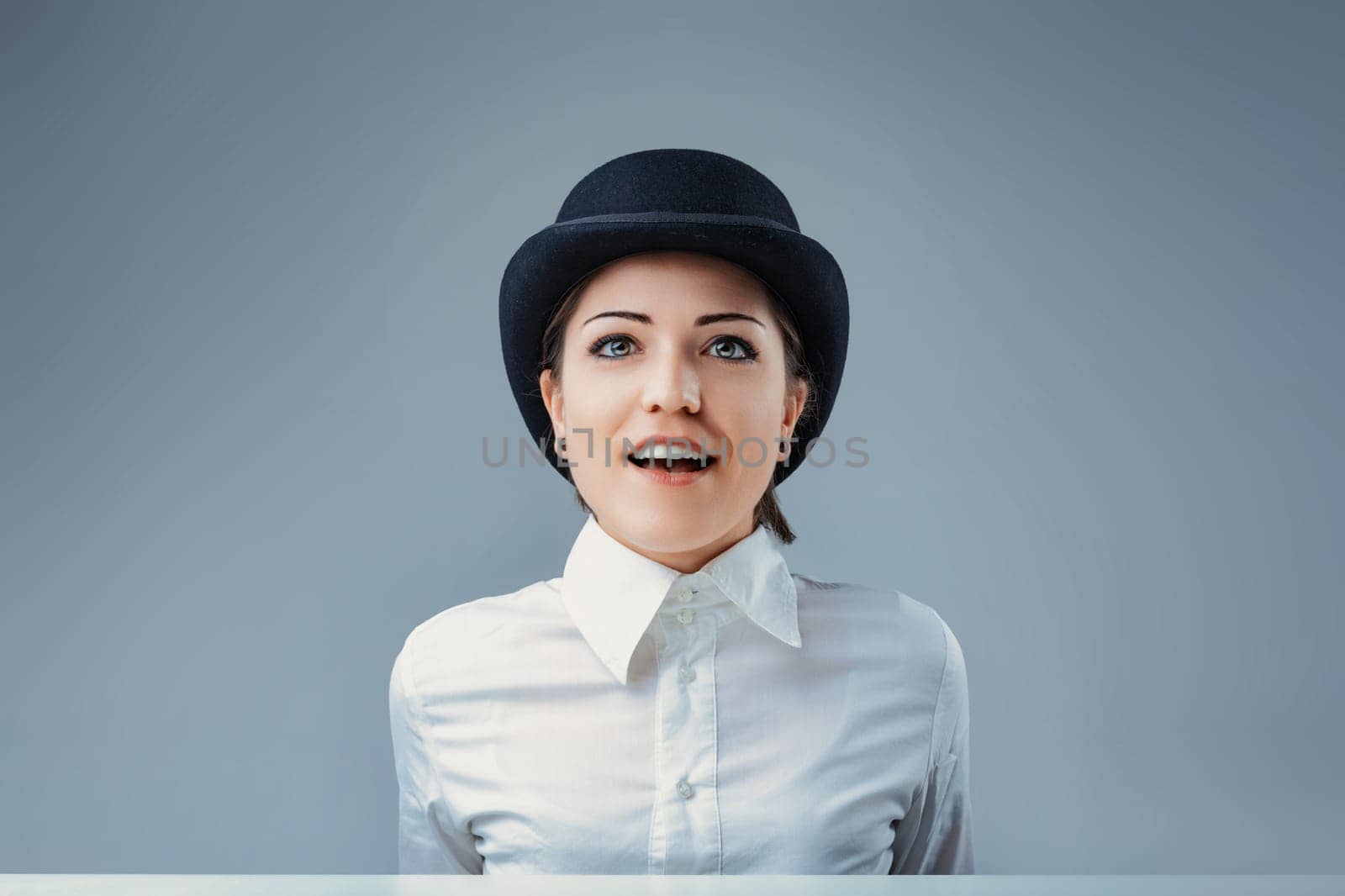 Portrait of a surprised young businesswoman looking up while wearing a bowler hat and a white shirt, isolated on a gray background, expressing amazement and curiosity