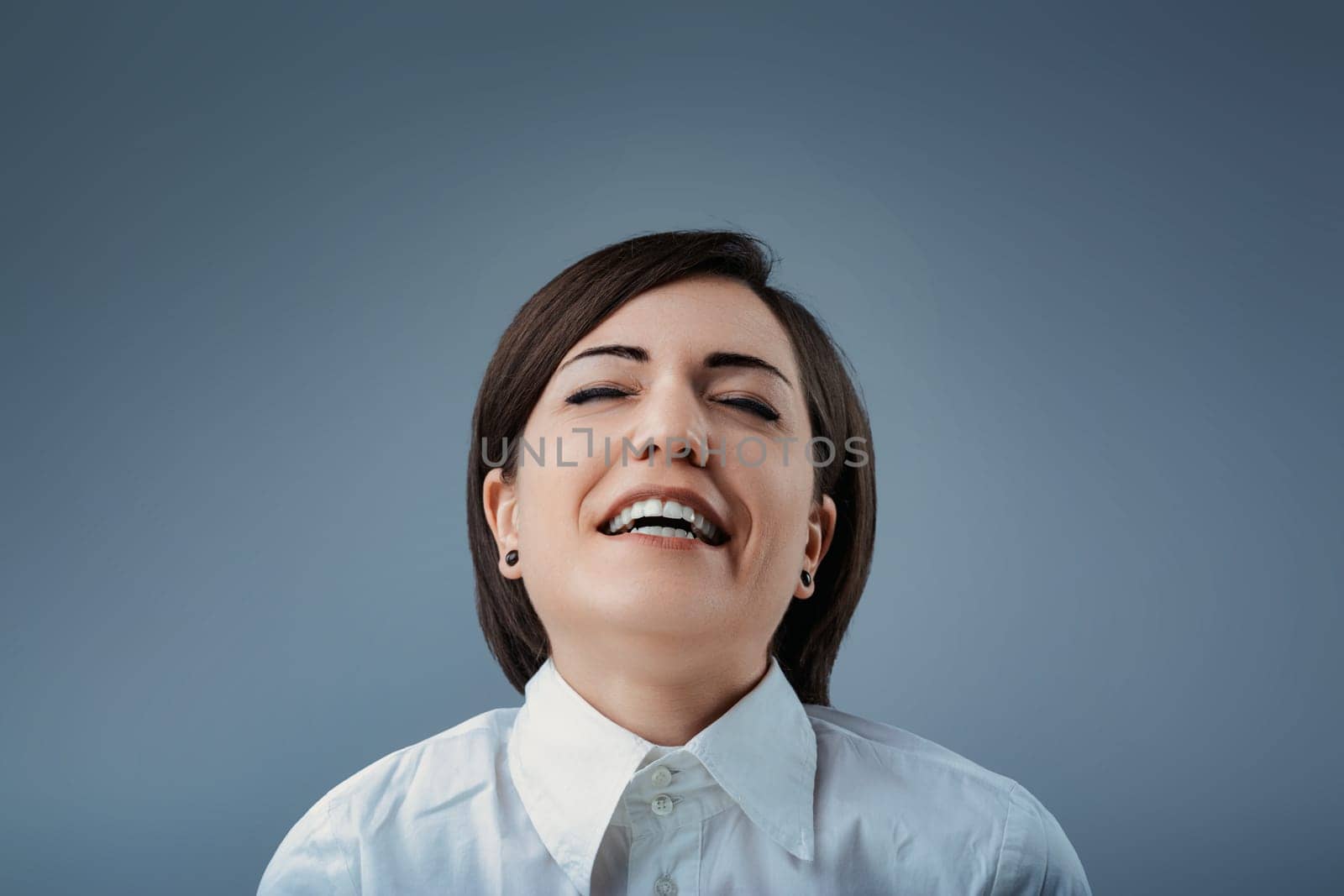 Studio portrait of a businesswoman laughing with her eyes closed, wearing a white shirt against a blue-gray background, expressing joy and happiness
