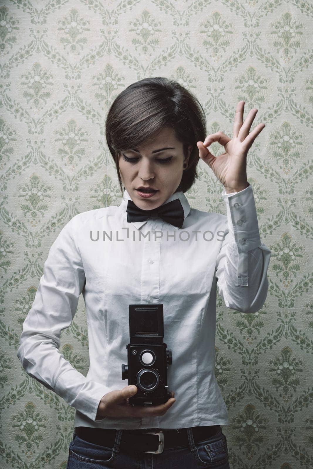 Young female photographer wearing a bow tie and holding a vintage twin-lens reflex camera gestures okay with her hand in front of a patterned wallpaper background