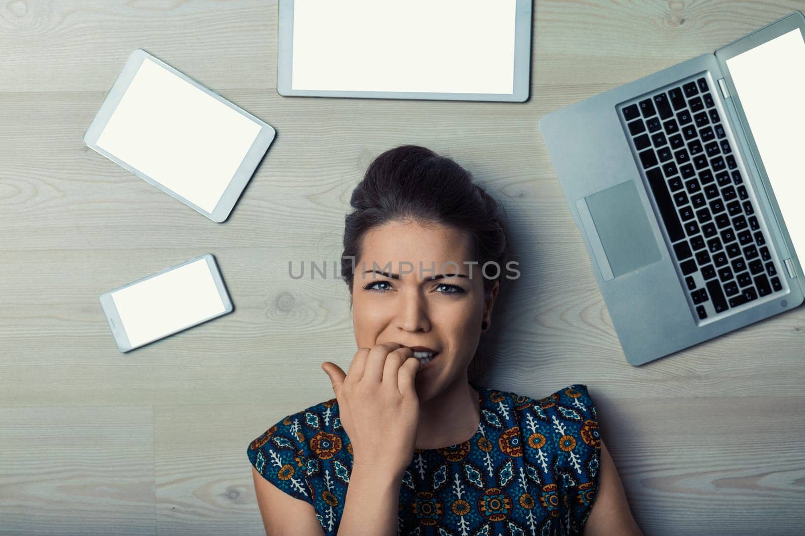 Stressed young woman is lying on the floor, surrounded by a laptop, tablet, and phone, biting her nails with a worried expression