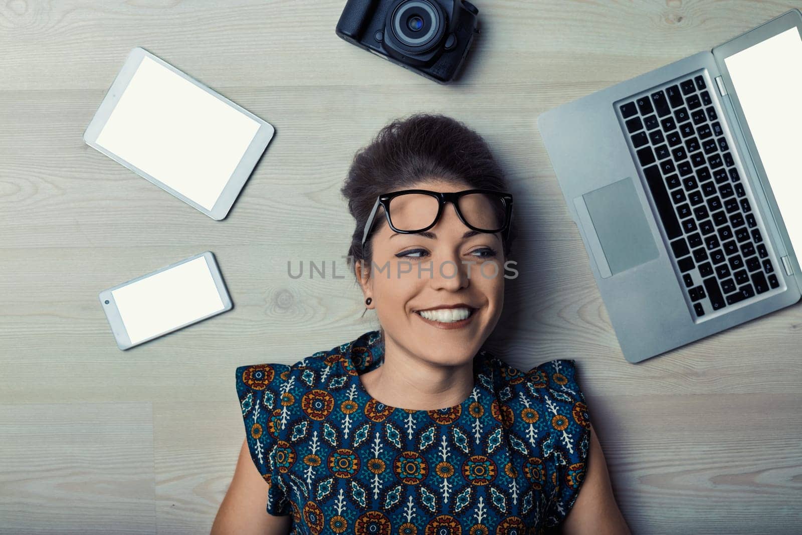 Smiling young woman lying on the floor with eyeglasses slightly down her nose, surrounded by several devices with blank white screens, looking happy and relaxed