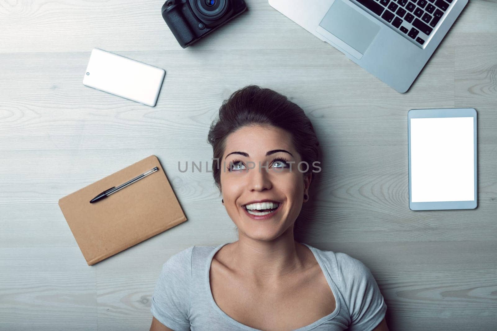 Young woman is lying on a wooden floor, smiling and looking up, surrounded by various technological devices, including a laptop, tablet, smartphone, camera, and notebook