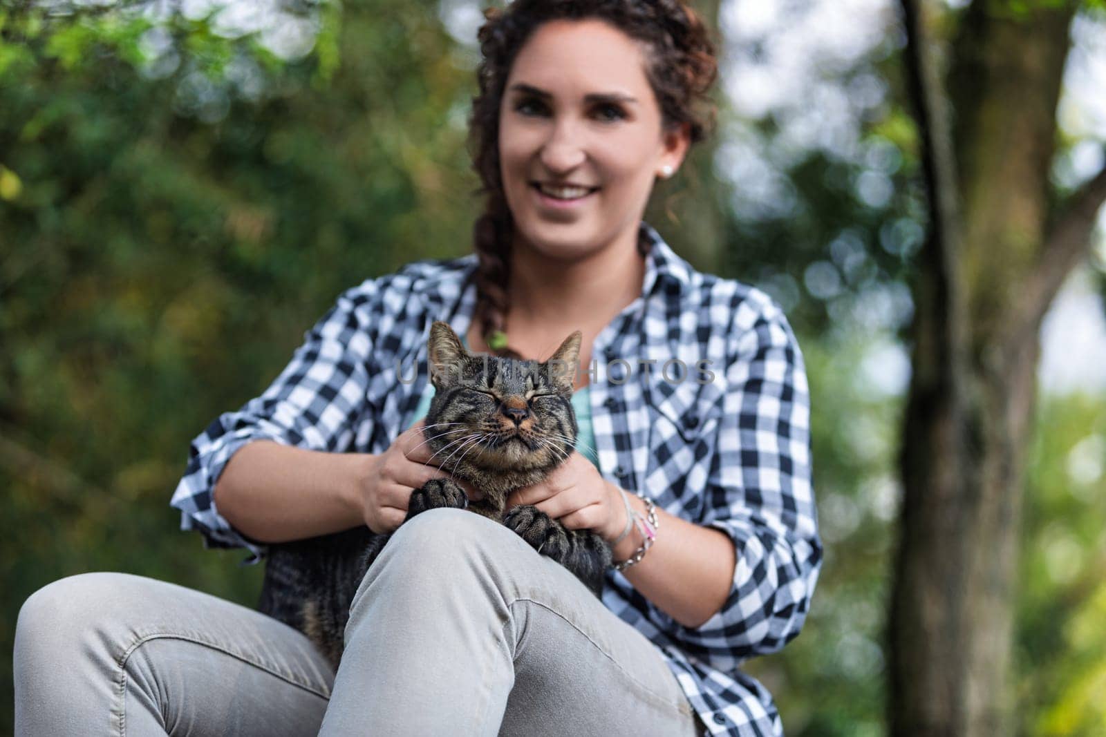 Young woman sits peacefully in a lush forest, holding a tabby cat on her legs, exuding love and care